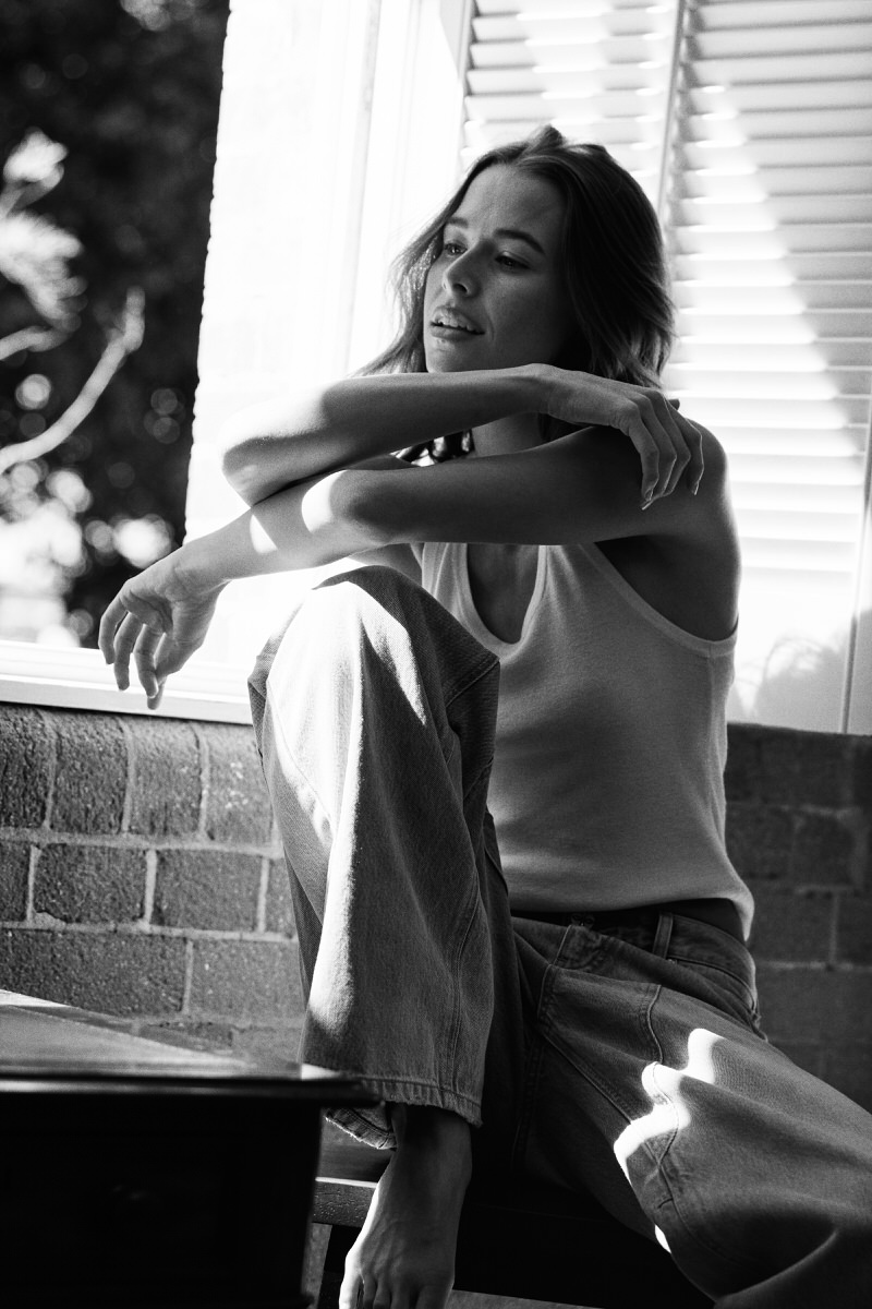 Jess Holiday seated beside a bright louvred window in a white tank top and jeans