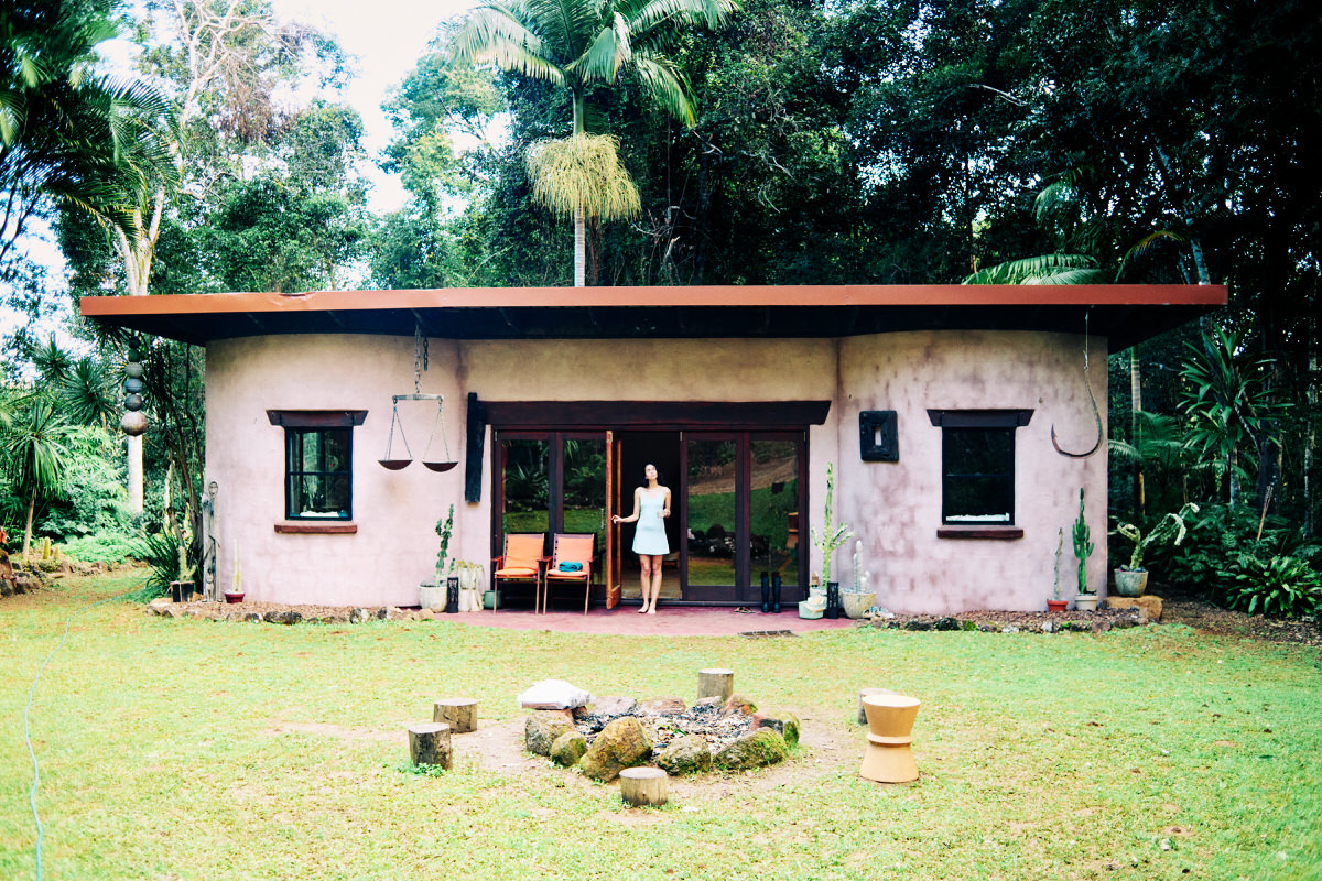 A small rendered house surrounded by a green subtropical garden, with Naomi standing in the open doorway