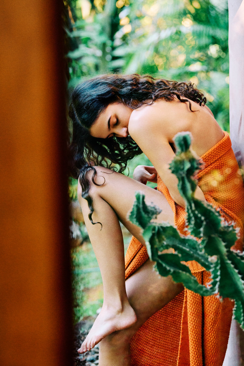 Naomi curled beside a cactus in bright orange knitted fabric, looking towards the ground