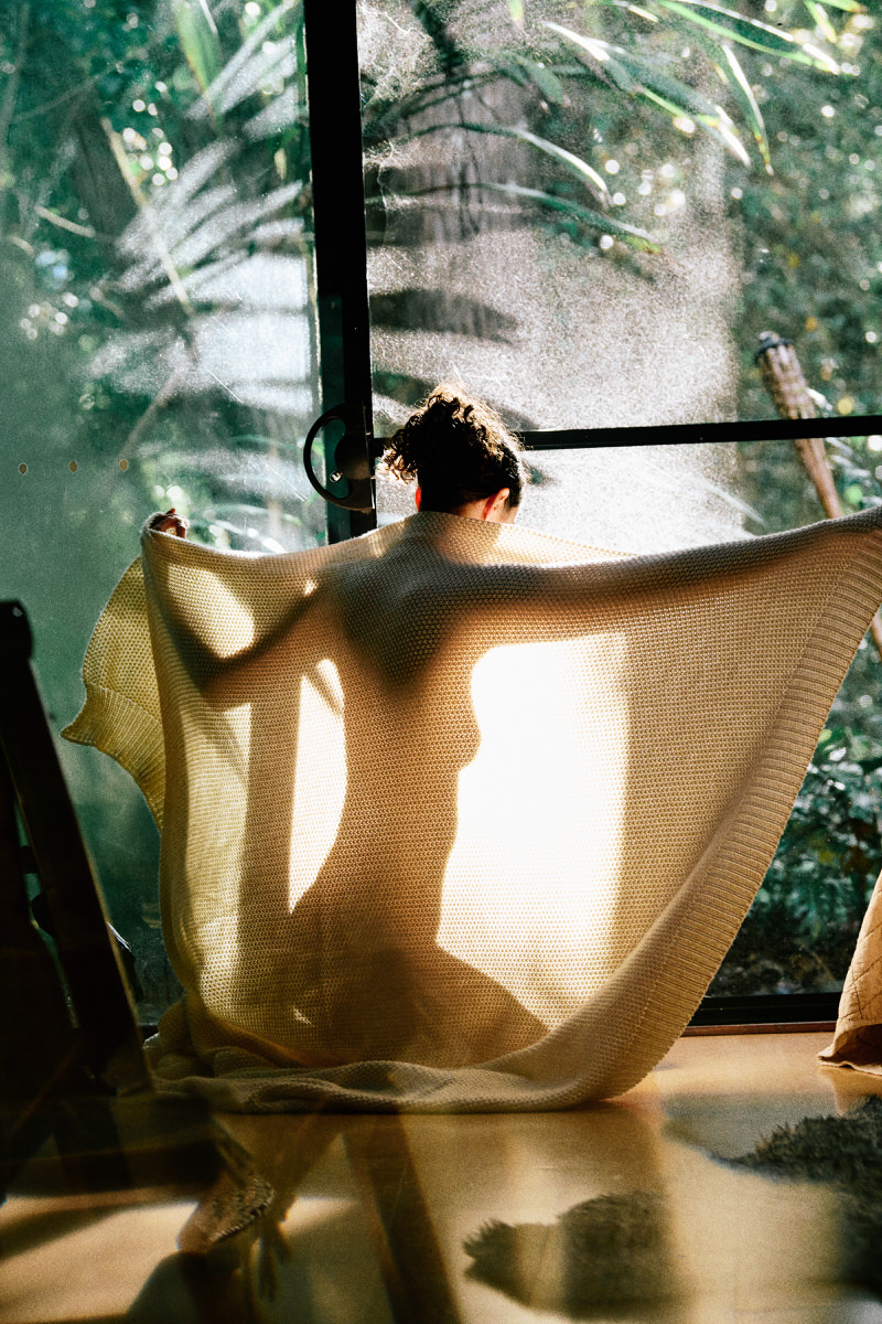 Seen from behind, Naomi holds a pale knitted blanket across her shoulders at a sunlit window