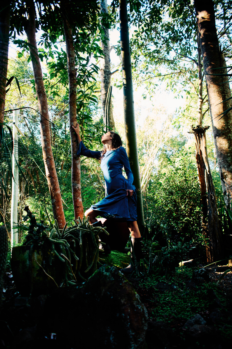 Naomi moves through dense subtropical trees in a blue dress, looking up into the canopy