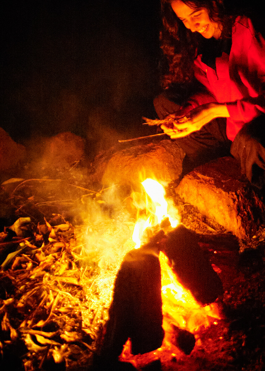Naomi crouches beside a small outdoor fire at night, lit by deep red and orange flames