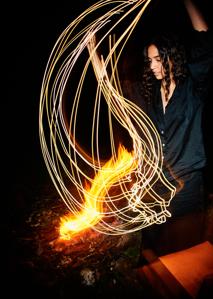 Naomi stands beside the fire as long trails of warm light arc through the dark frame