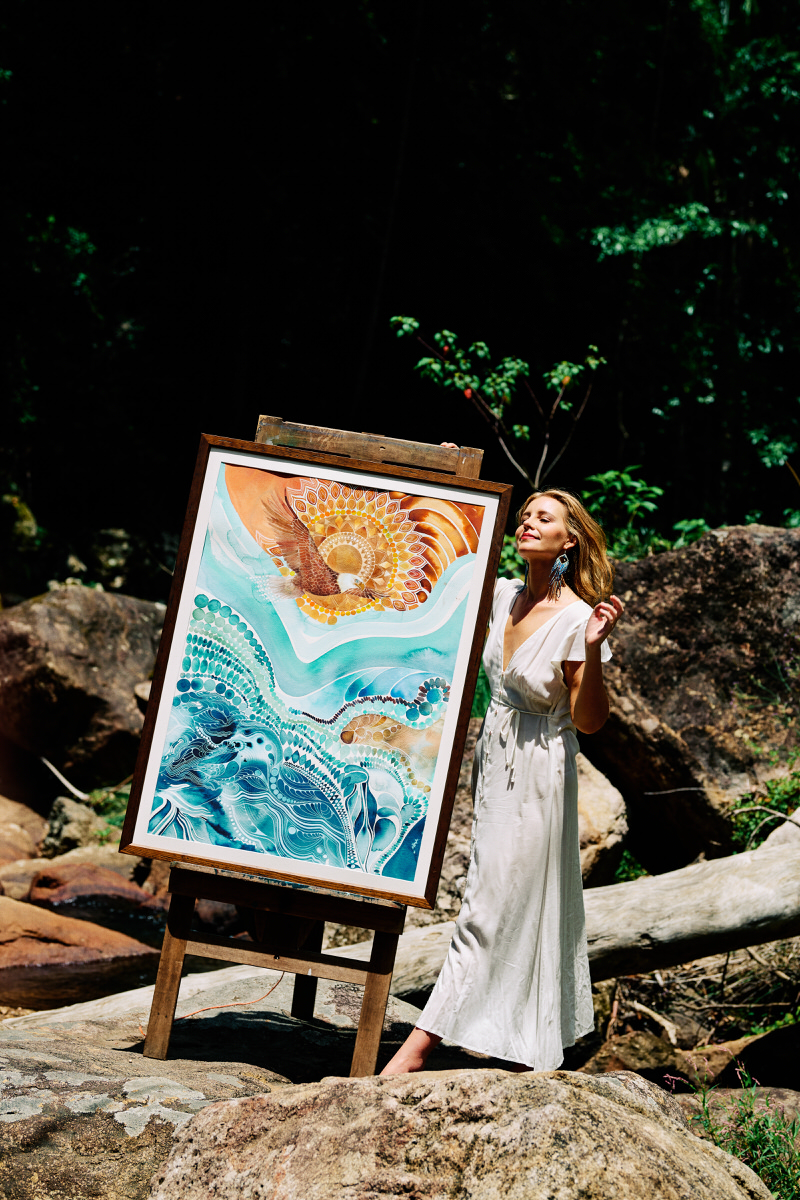 Christine standing beside her colourful painting on an easel among rocks and rainforest near Byron Bay